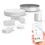 Éléments du kit Somfy Home Alarm Essential disposés à plat : sirène intérieure, boîtier Link Essential, deux détecteurs d'ouverture, clavier à code, détecteur de mouvement et capteur de vibration, à côté d'un smartphone affichant l'application avec le statut « Alarme désactivée »