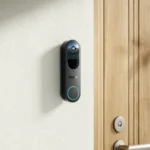 Sonnette vidéo Reolink Doorbell Battery fixée au mur près d'une porte en bois clair, voyant bleu allumé